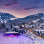 A photo taken of Whistler Village in the winter time in the purple hues of the evening alpen glow. Whistler and Blackcomb mountains are in the background with the festive lights of the village and Olympic Plaza in the foreground.