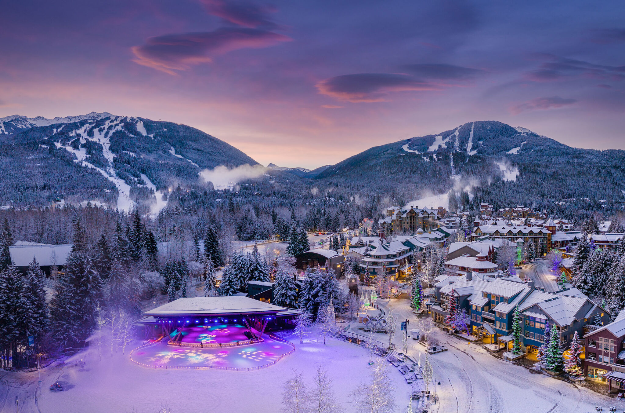 A photo taken of Whistler Village in the winter time in the purple hues of the evening alpen glow. Whistler and Blackcomb mountains are in the background with the festive lights of the village and Olympic Plaza in the foreground.