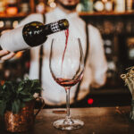 A server pours a glass of red wine, which is sat on a wooden bar.