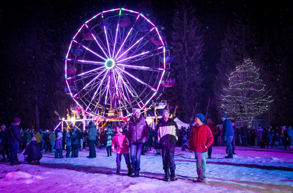 A family enjoys the Winter Light celebration in Whistler. A large, brightly lit Ferris wheel glows in the snow behind them. 