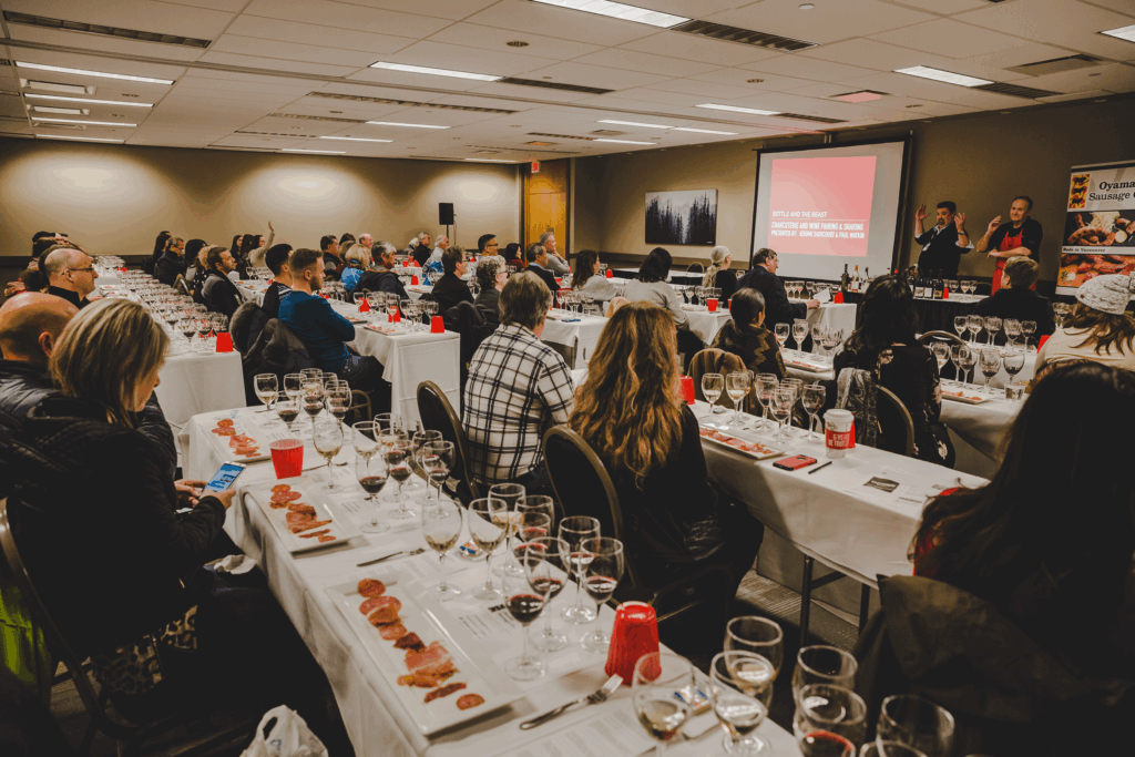 A room full of people seated at desks with two presenters at the front. A selection of wine and meats are laid out in front of each person. 