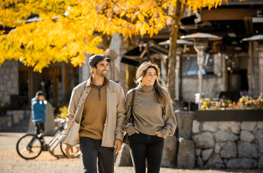 Two people walk down Whistler Village Stroll, a beautiful tree is turning yellow behind them showing off its fall colours.