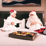 Santa and Mrs. Claus sit on a plush white bed at the Fairmont Chateau Whistler dressed in white spa robes. A platter of nibbles are on a tray in front of them.