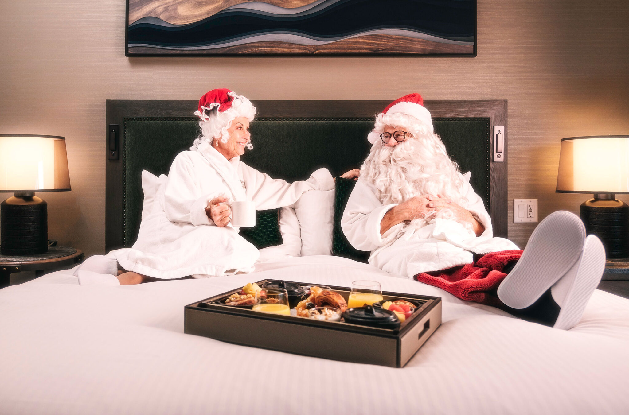 Santa and Mrs. Claus sit on a plush white bed at the Fairmont Chateau Whistler dressed in white spa robes. A platter of nibbles are on a tray in front of them.