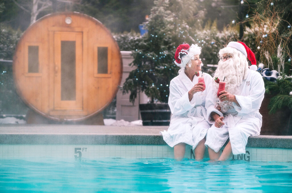 Santa and Mrs. Claus sit on the side of the pool at the Fairmont Whistler drinking smoothies with the barrel sauna in the background.