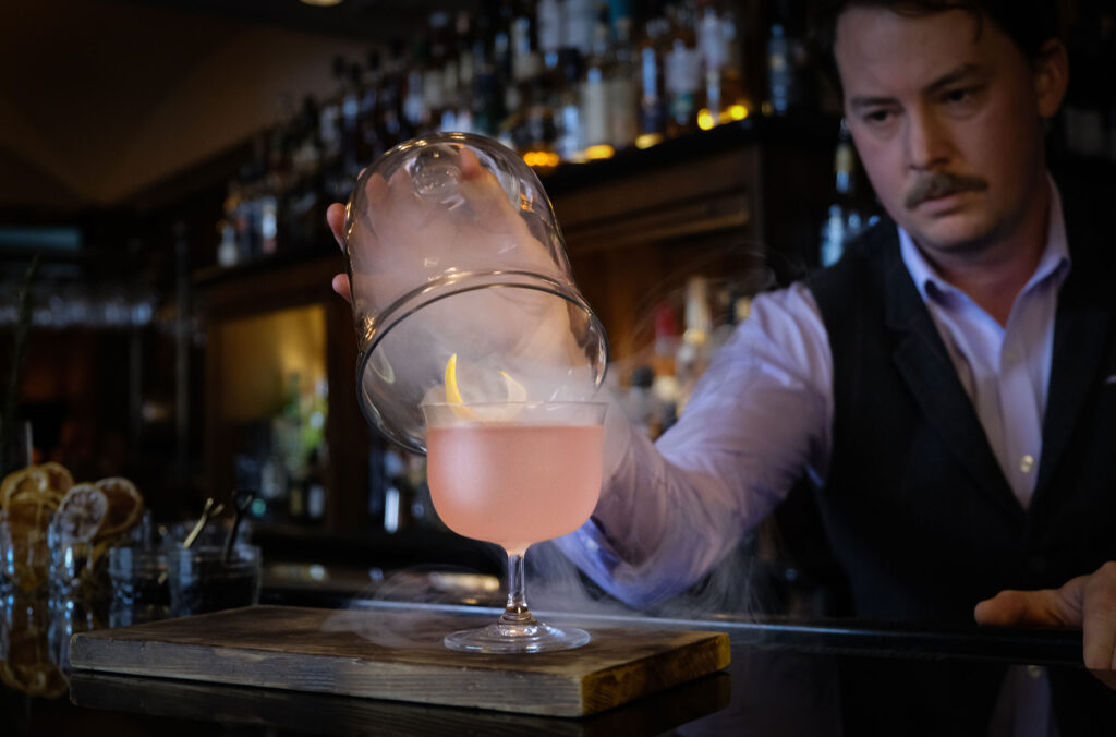 A bartender takes a glass covering off a candy-pink cocktail and smoke billows out across the bar at The Mallard Lounge at Fairmont Chateau Whistler.