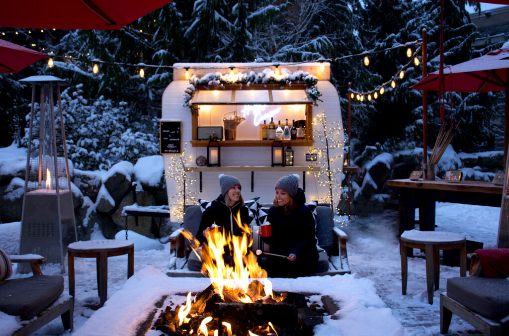 Two people sit on the Four Season's inner courtyard patio which is covered in snow. A small, vintage camper is in the background, and is where drinks are being served from. They're happily drinking hot chocolate and toasting marshmallows over a fire.