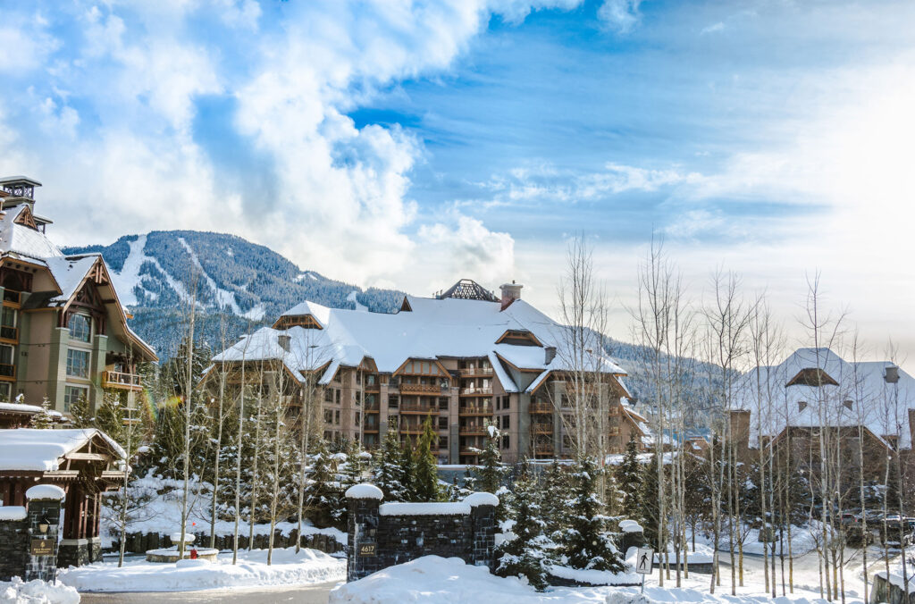 A shot of the Four Seasons Resort & Residences Whistler in the winter. Snow blankets the hotel, with Whistler Blackcomb in the background.