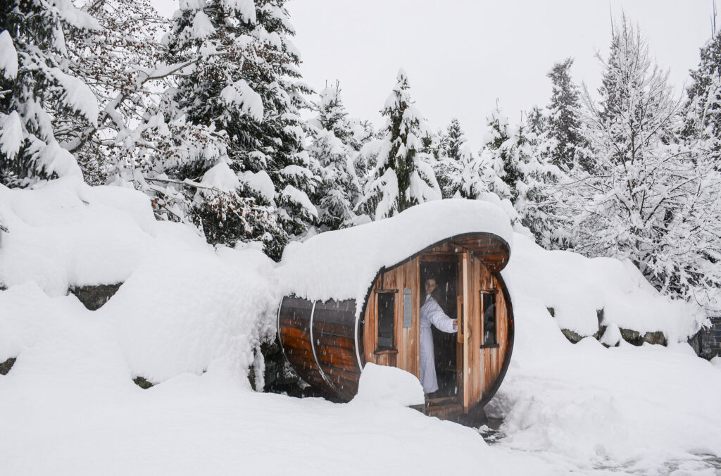 A person wearing a fluffy white robe emerges from a snow-laden barrel sauna sat on the pool deck at the Four Seasons Whistler.