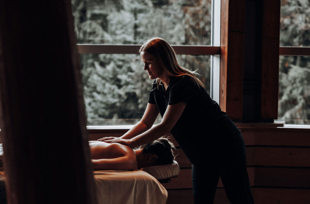 A massage therapist massages a person laying on a massage table in the spa at the Four Seasons Whistler. You can see the forest out the windows in the back of the shot.