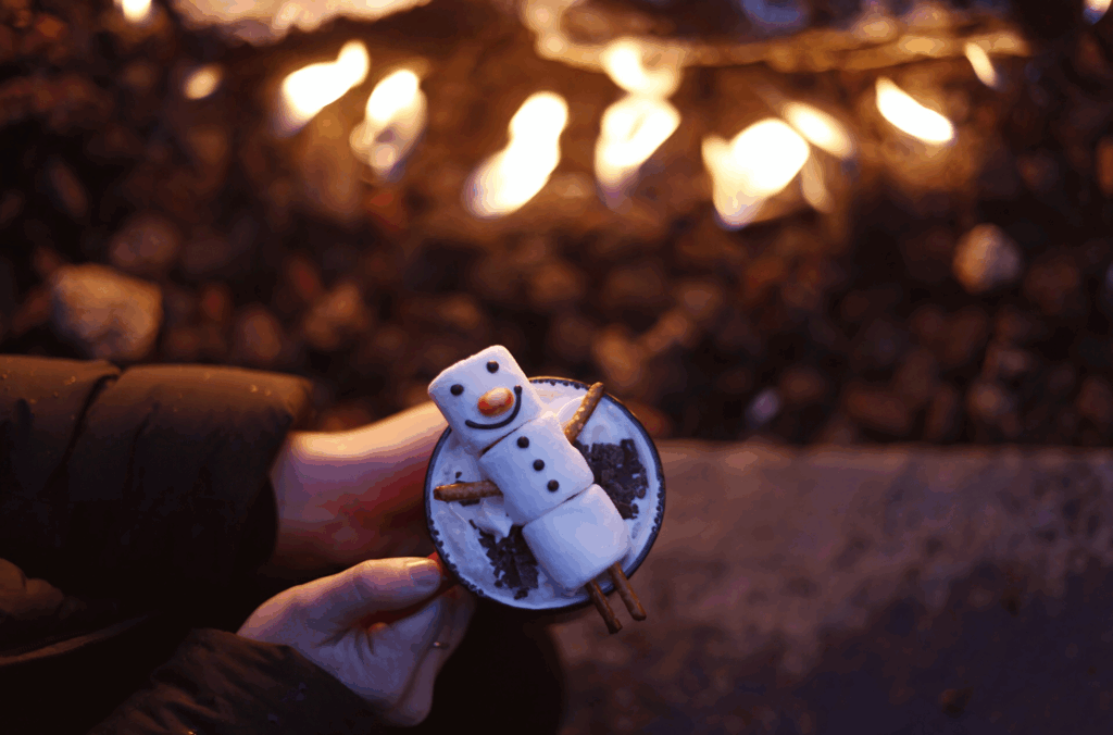 A person holds a mug with a tipsy snowman balancing on top. His marshmallow body will slowly dissolve into the hot chocolate below. They are by an outdoor fire on the patio of the Four Seasons Whistler.