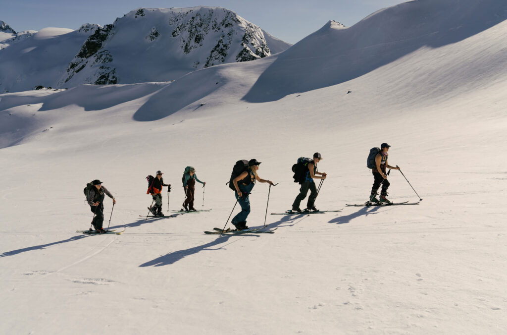 A group of six people on skis skin up the mountain in the Whistler backcountry in the sunshine. The skies are blue and the snow is crisp and white.