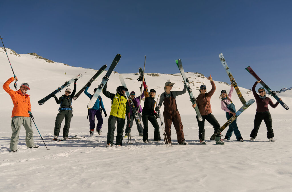 A group of people hold their skis up in the air cheering. They're on a women's camp with Extremely Canadian in Whistler's snowy backcountry.