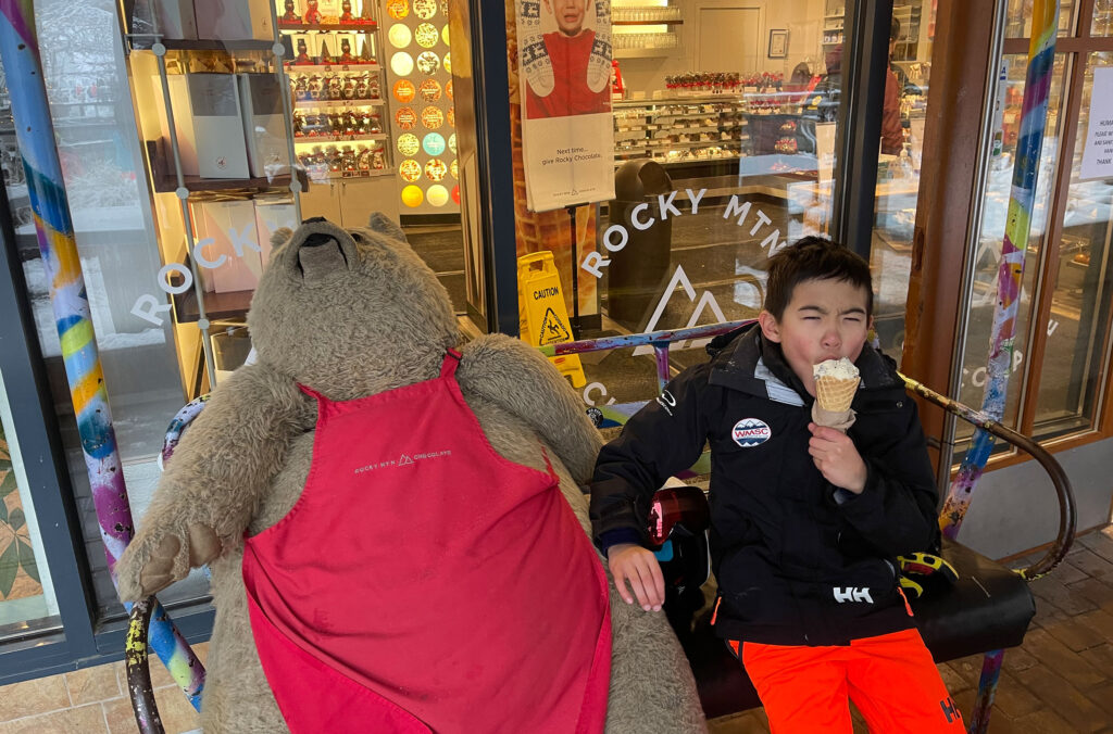 A young person dressed in ski gear eats an ice cream sitting next to a stuffed bear that resides outside of The Rocky Mountain Chocolate shop in Whistler. 