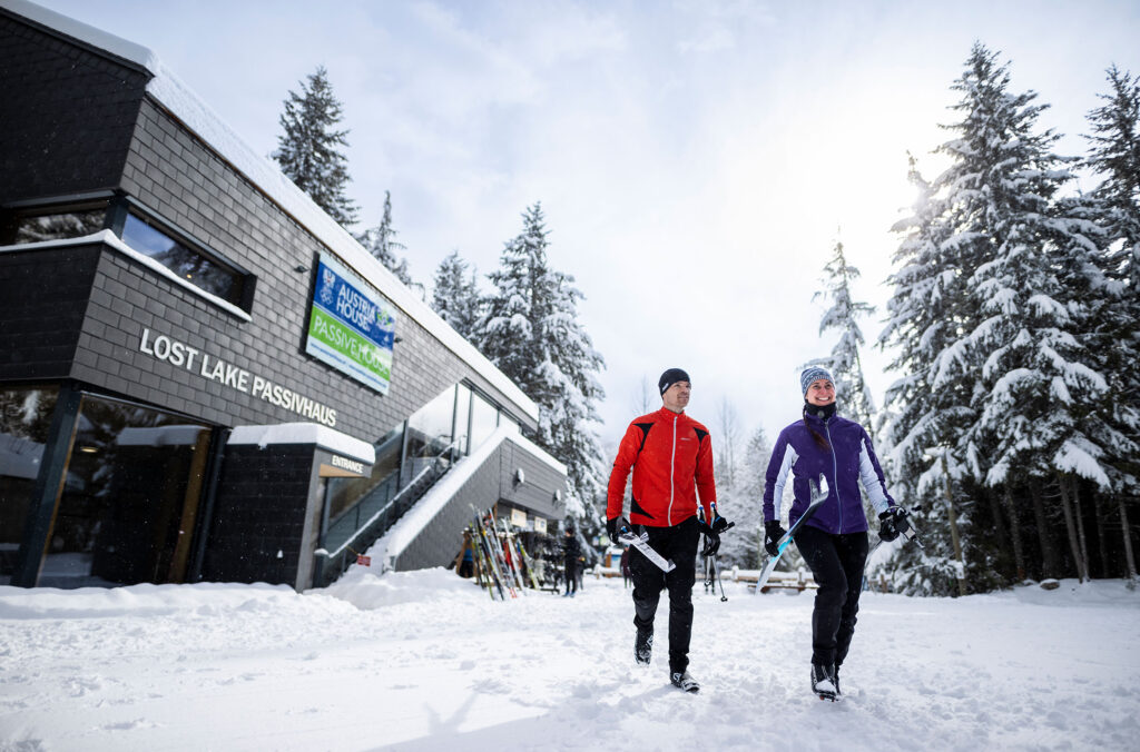 Two people carry cross-country ski gear as they exit the Lost Lake Passivhaus toward the trails.