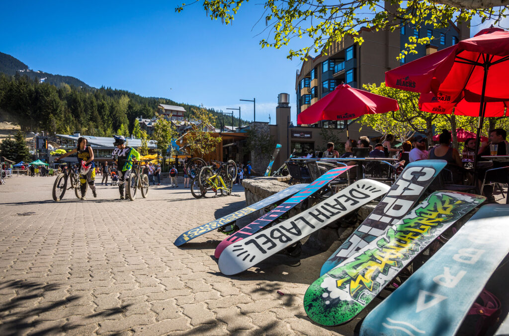 Whistler's Skier's Plaza in the spring sunshine. Bikers walk their bikes close to the bike park lift while snowboarders rest their boards while they enjoy the patio at Black's.