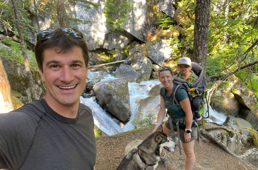 Liz Francis hikes with her family in Whistler in the summer. She carries her young child in a backpack and also holds her dog.