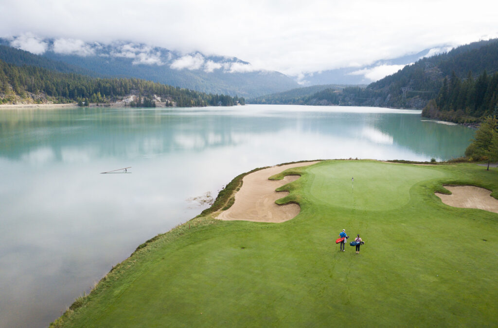 An aerial shot of the edge of Nicklaus North Golf Course on the edges of Green Lake with the mountains in the background in Whistler.