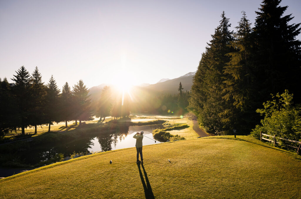 A golfer tees off in the early morning light, with the sun rising over the mountains and casting long shadows on the lush green fairway in Whistler.