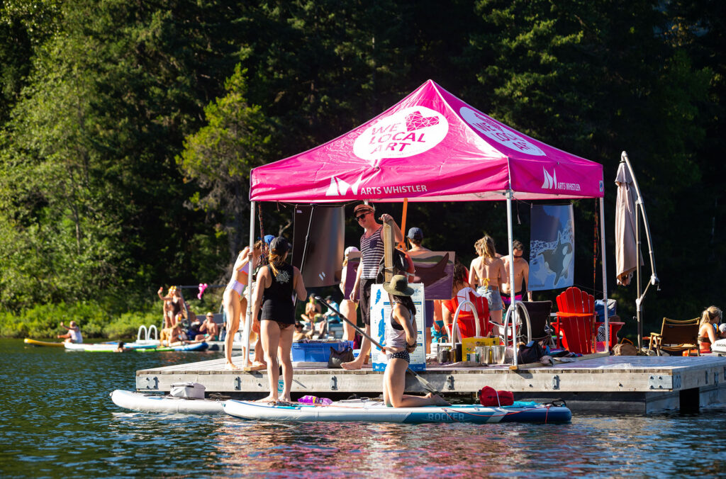Paddle boarders float next to a dock with artists painting under a pink awning.