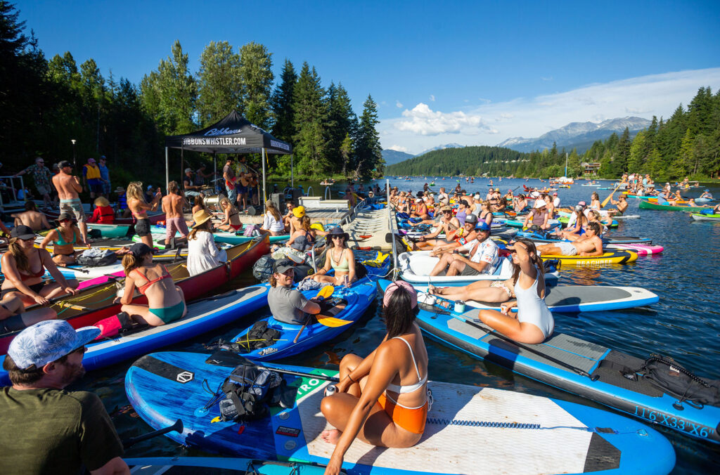 Paddle boarders and kayaks float next to a lake dock with musicians playing in the sunshine.
