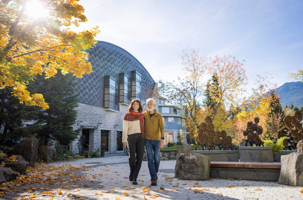 A couple walk through Whistler Village in the fall. Golden trees line the walkway as they stroll past a piece of public art.