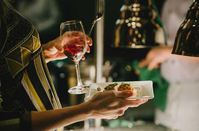 A person holds a glass of wine while reaching for food at a food station at the Night Market event at Whistler Cornucopia.