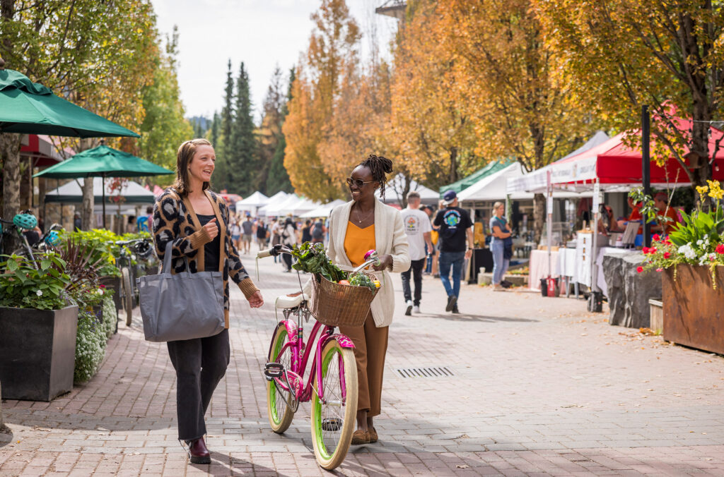 Two people walk down Whistler's Upper Village during the Whistler Farmers' Market. One has a bike loaded with fresh produce and the other a large bag. The trees are yellow and gold behind them, with market stalls in front.