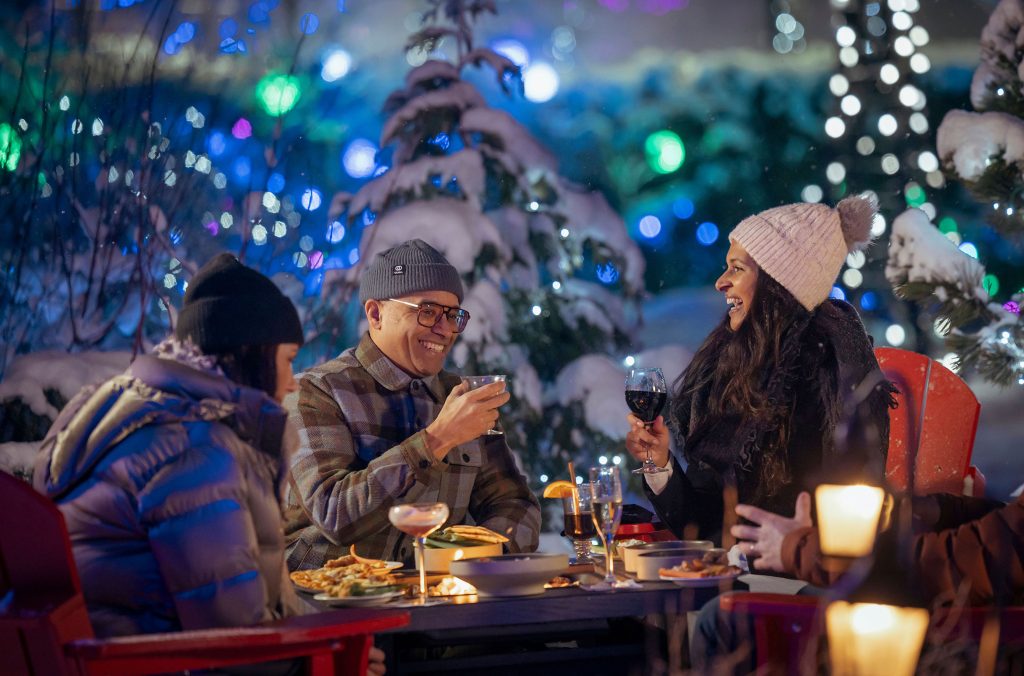 Friends enjoy drinks on an outdoor patio lit by festive lights in Whistler Village.