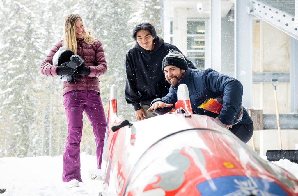 A guide stands next to a red bobsleigh at Whistler Sliding Centre explaining what happens during a bobsleigh experience to two people.
