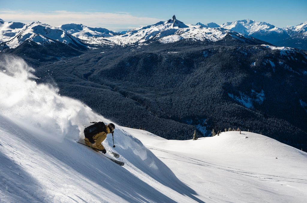 A skier sends a up a cloud of snowy powder behind them as they make their way down a slope on Whistler Blackcomb with Black Tusk in the background.