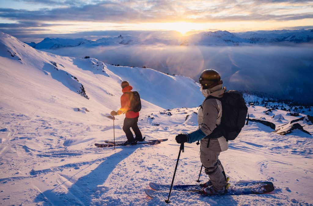 Two skiers stop at the top of a trail on Blackcomb Mountain as the sun sets behind the Coast Mountains.