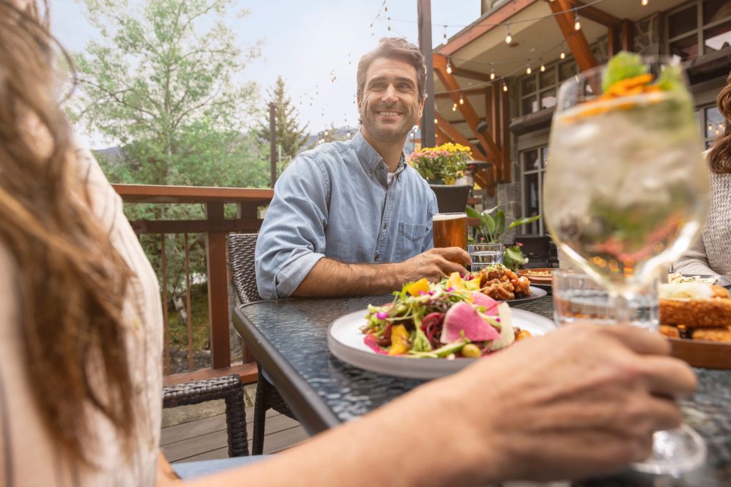A group of people dine outside on a Whistler patio in the spring. Plates of colourful food are laid on the table and fairy lights hang above.