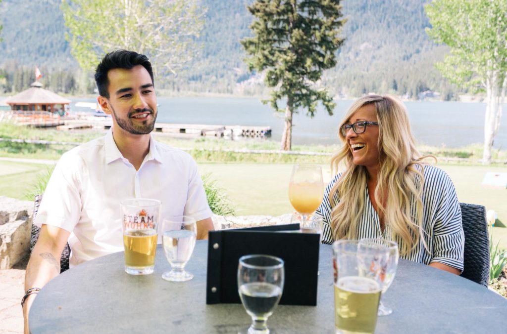 A couple sit at an outdoor table at Table Nineteen in Whistler, overlooking Green Lake in the summer.