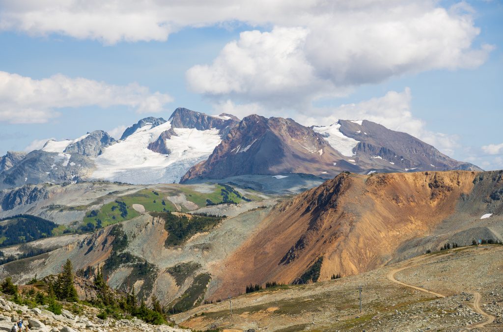Expansive alpine view from Whistler featuring Fissile Peak in the distance, with red-orange ridgelines, grassy meadows, and glacier-covered peaks.