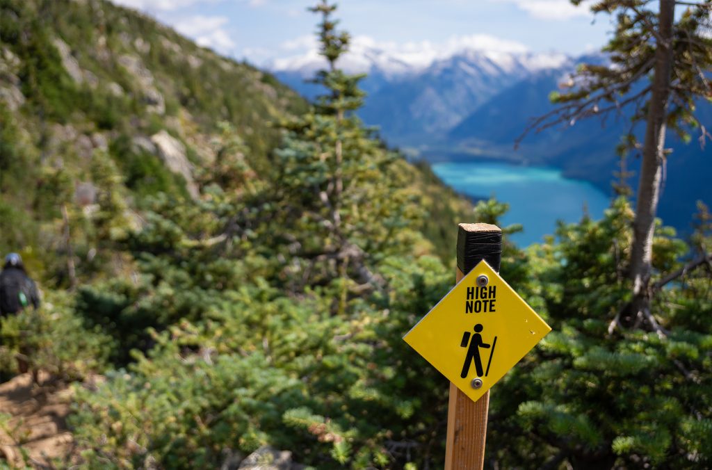Yellow trail marker for the High Note Trail set against alpine greenery, with Cheakamus Lake’s vibrant blue visible in the distance - an early waypoint on the route to the Kees and Claire Hut.