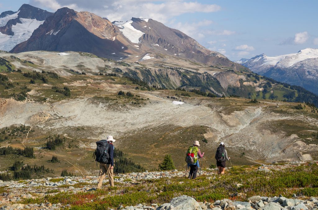 Three hikers making their way along the Musical Bumps Trail toward Russet Lake, surrounded by glaciers, rocky peaks, and alpine meadows.