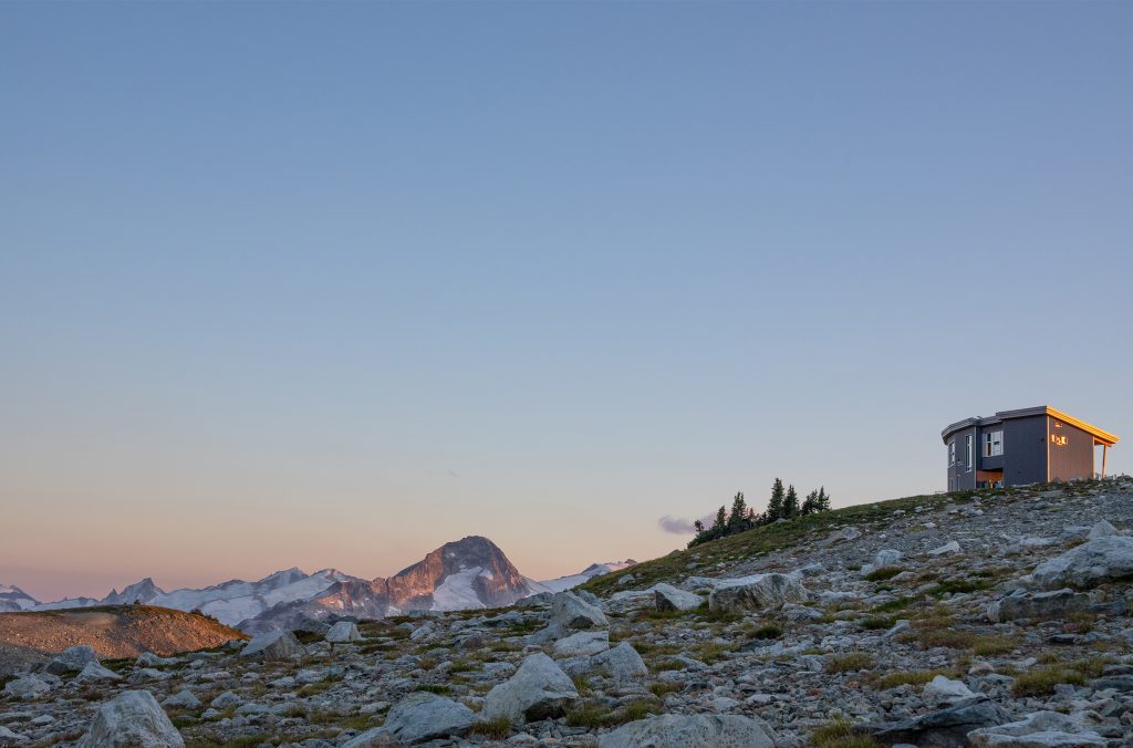 Kees and Claire hut glowing in the sunset, perched high in the alpine, with peaks in the distance.