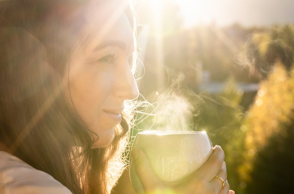 A person drinks from a steaming cup outside with the sun shining on them.