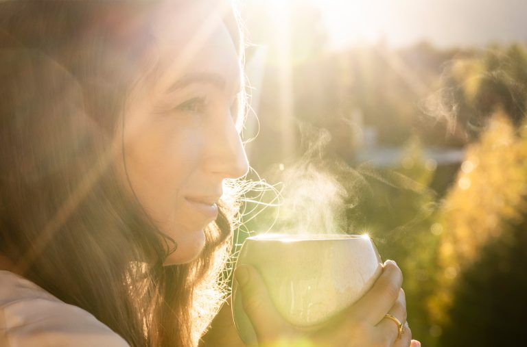 A person drinks from a steaming cup outside with the sun shining on them.