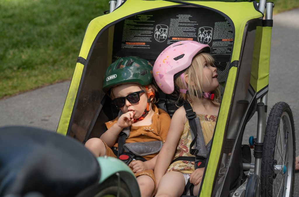 Two toddlers wearing helmets in a chariot that attaches to a bike.