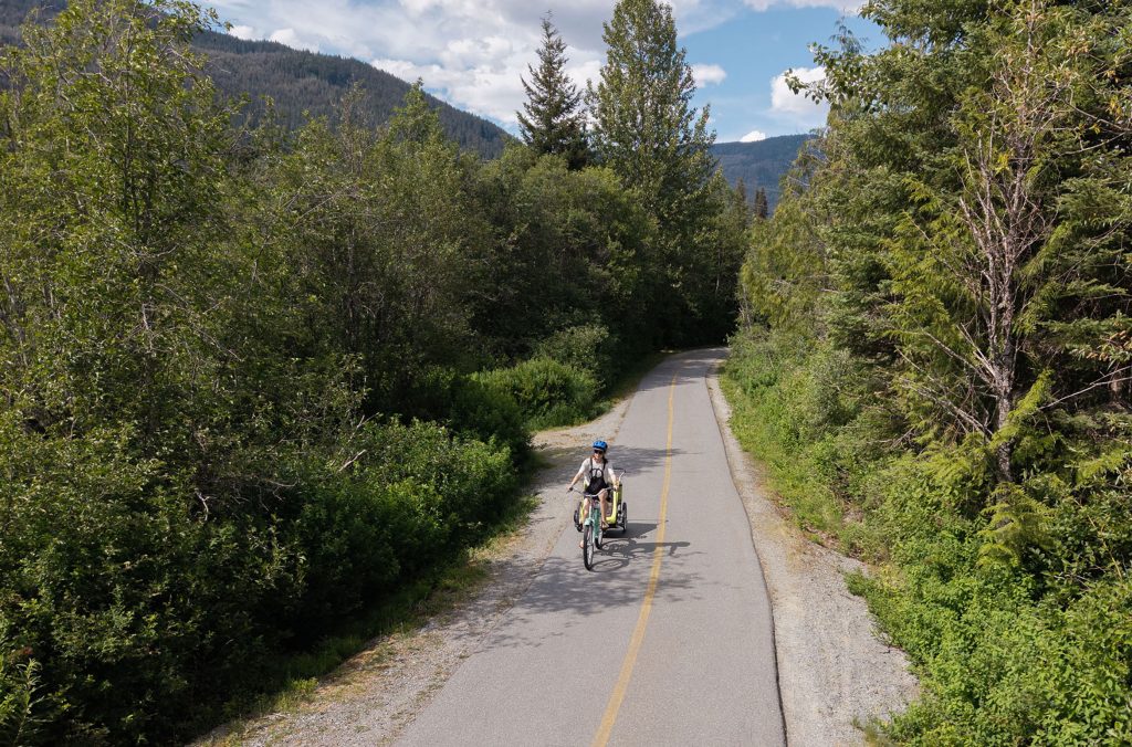 A biker rides along the Whistler Valley Trail pulling toddlers in a chariot. Lush evergreens are on all sides of the path and it's the summer.