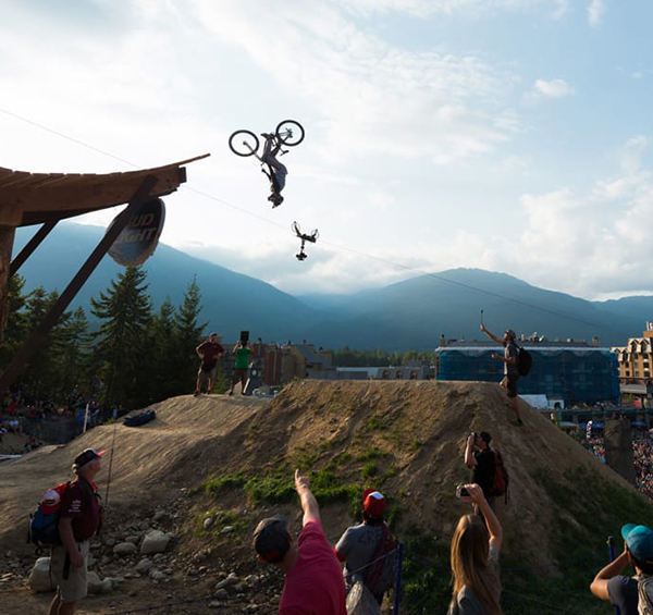 A mountain biker soars upside-down in a daring aerial trick during Crankworx Whistler bike festival, with a crowd watching against a stunning alpine backdrop. The energy is high, the stoke is real.