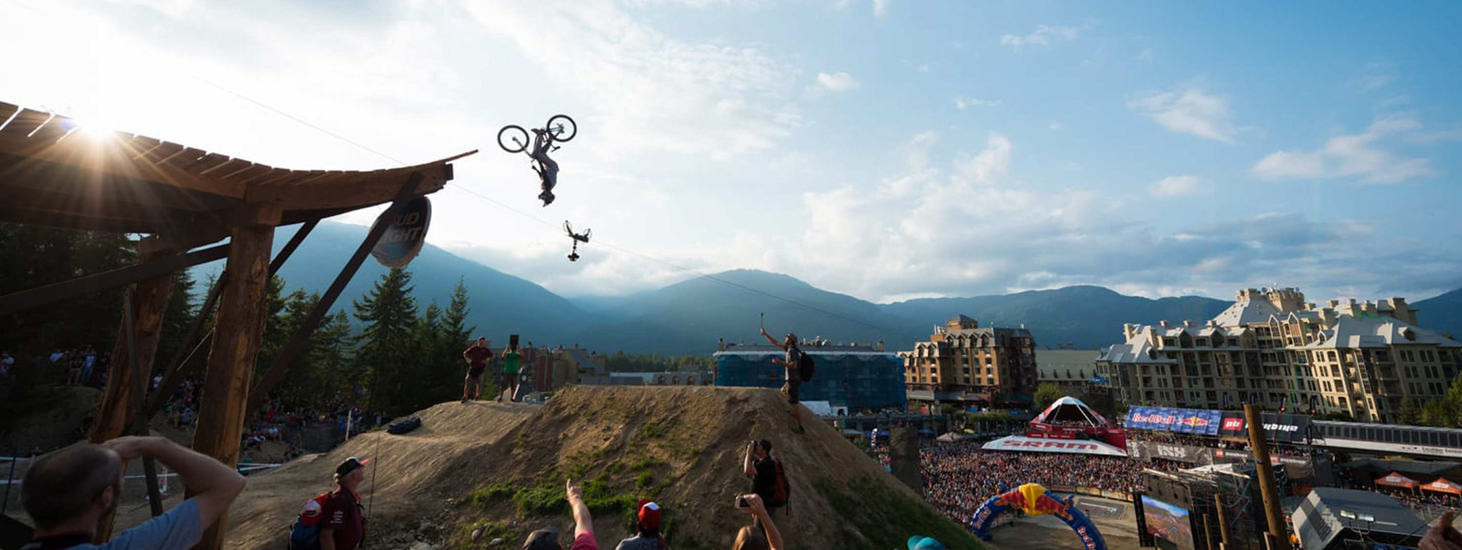 A mountain biker soars upside-down in a daring aerial trick during Crankworx Whistler bike festival, with a crowd watching against a stunning alpine backdrop. The energy is high, the stoke is real.