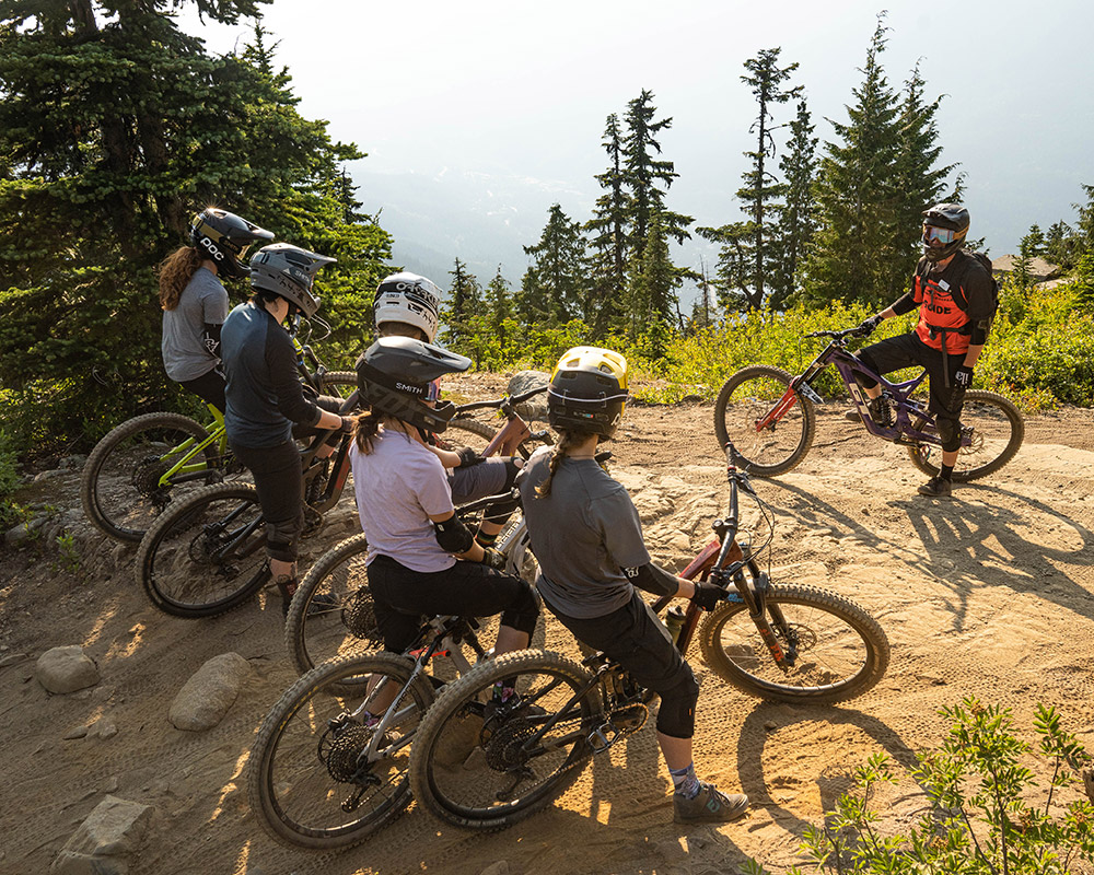 A group of mountain bikers, clad in full protective gear and helmets, pause on a rugged trail in Whistler, listening to their guide. Framed by towering evergreens and a sunlit alpine backdrop, the riders take in expert advice before dropping into their next thrilling descent.