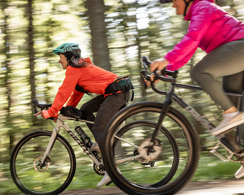 Two cyclists cruise through a sun-dappled forest trail in Whistler, enjoying the smooth ride of high-quality rental bikes. From downhill rigs to trail-ready enduro bikes, there’s a perfect ride for every adventure.