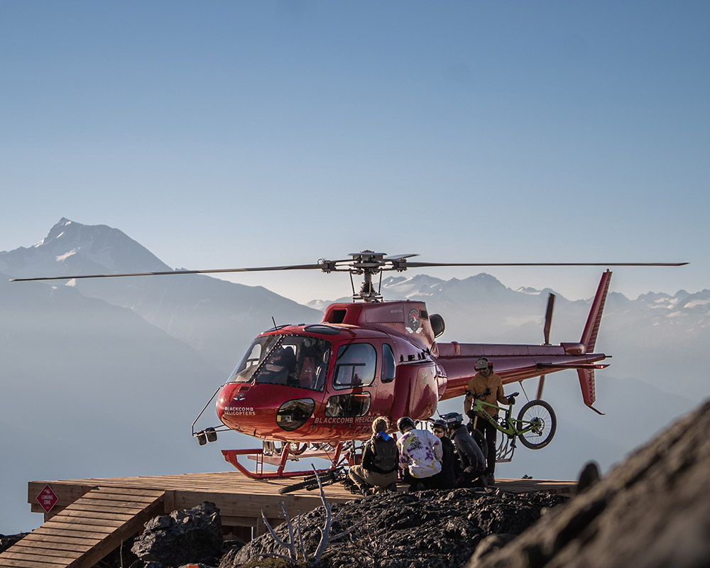 A red helicopter lands on a remote mountain ridge in the Coast Mountains, unloading riders and their bikes for an epic heli-biking adventure. Rugged terrain, alpine singletrack, and breathtaking views await.