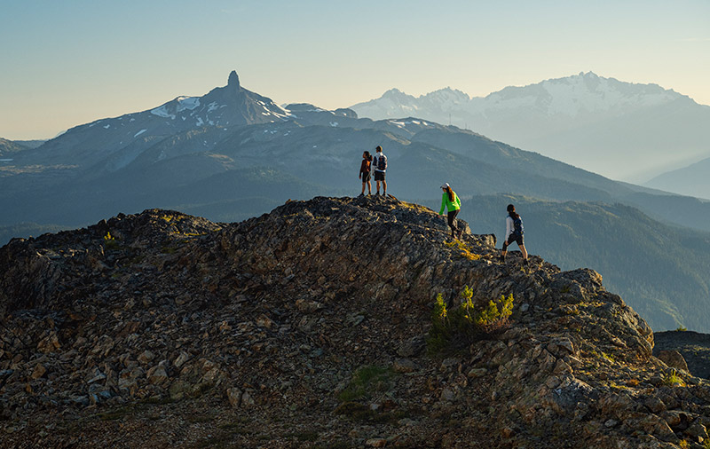 Hikers stand atop a rugged mountain ridge in Whistler, soaking in panoramic views of jagged peaks, including the iconic Black Tusk. The alpine air is crisp, the adventure limitless.