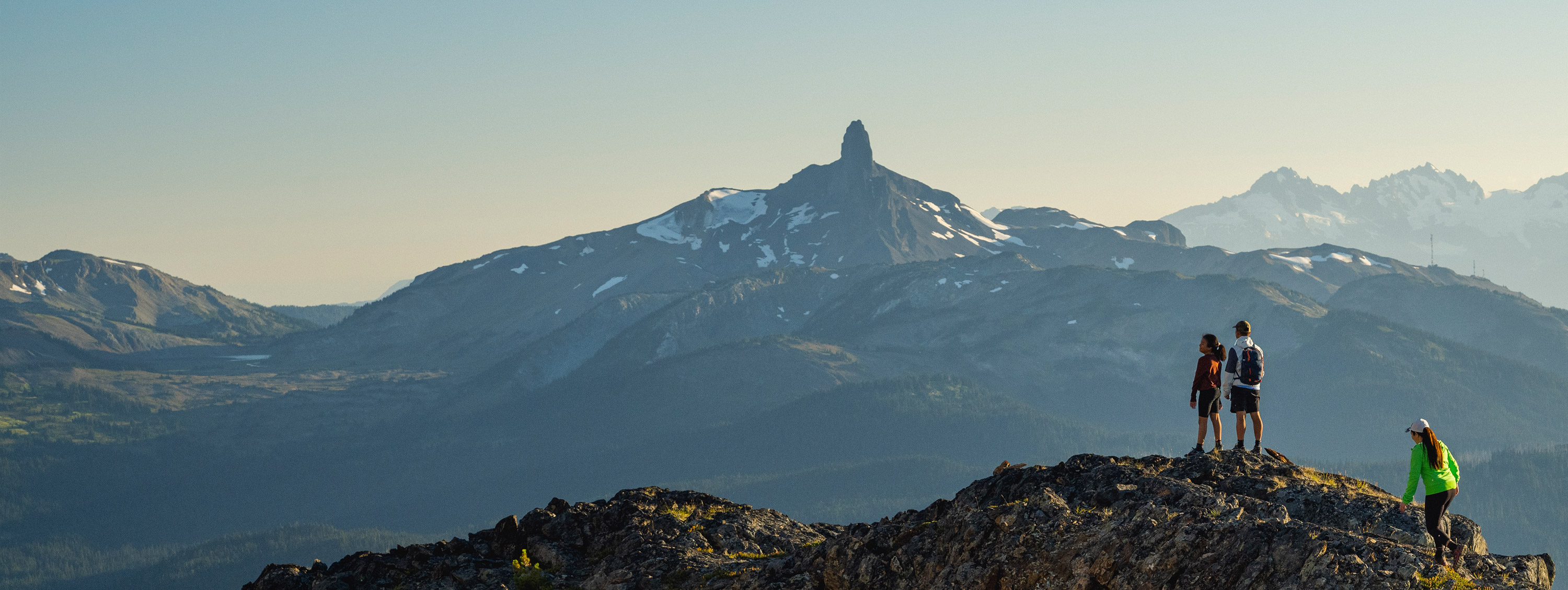 Hikers stand atop a rugged mountain ridge in Whistler, soaking in panoramic views of jagged peaks, including the iconic Black Tusk. The alpine air is crisp, the adventure limitless.