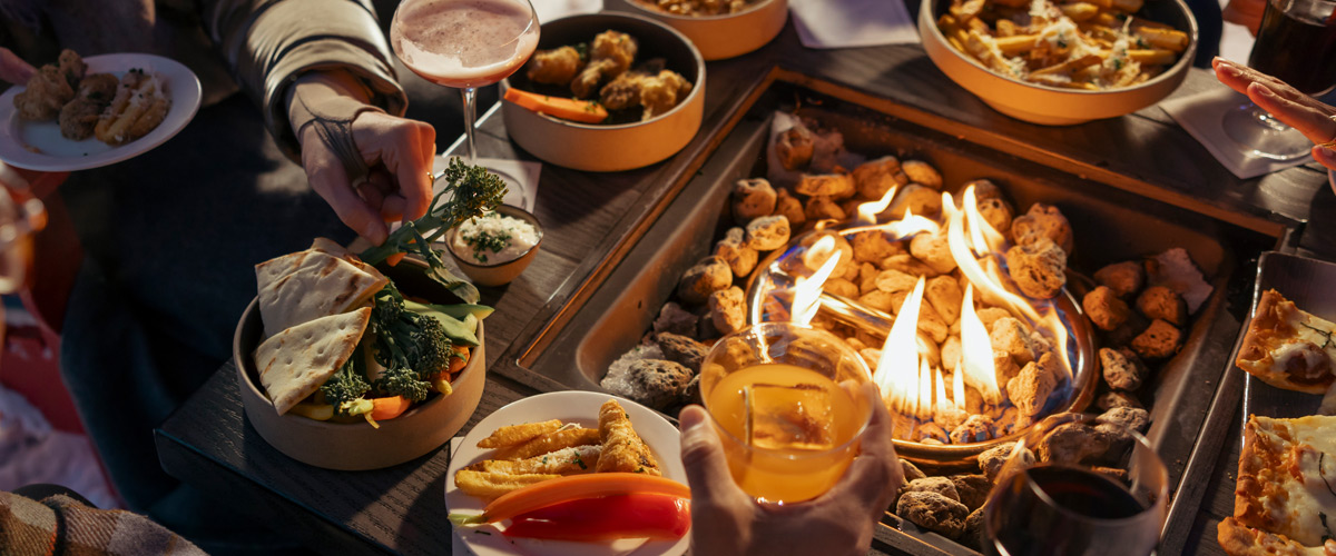 Group of people gathered around an outdoor fire table enjoying shared plates of food and drinks in Whistler, BC